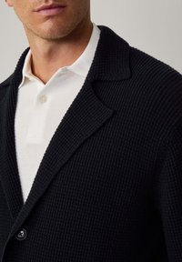 Svart texturerad stickad cardigan med bred krage, enknappsstängning, över en vit skjorta med krage. Närbild på detaljerna kring överkroppen.