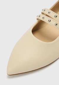 Beežid terava ninaga jalanõud, sileda nahkviimistlusega, millel on astmikunaha kohal kaks dekoratiivset rihma metallist naelastega.