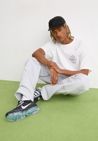 White t-shirt with a logo, gray baggy pants, and black sneakers with a translucent sole. The individual is wearing a black cap and sitting on green flooring.