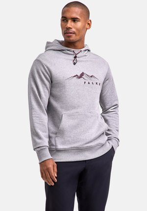 Uomo con felpa con cappuccio grigio chiaro con grafica di montagna e scritta "FALKE", abbinata a pantaloni scuri, in piedi contro uno sfondo neutro.