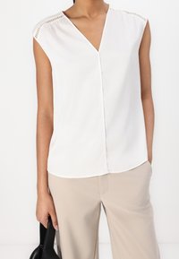 Blouse blanche sans manches avec un col en V, ornée de coutures décoratives sur les épaules. Associée à un pantalon beige et un sac noir.