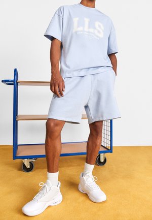 Person wearing light blue LLS shorts and matching T-shirt, white socks, and white sneakers standing near a blue and wood cart on yellow floor.