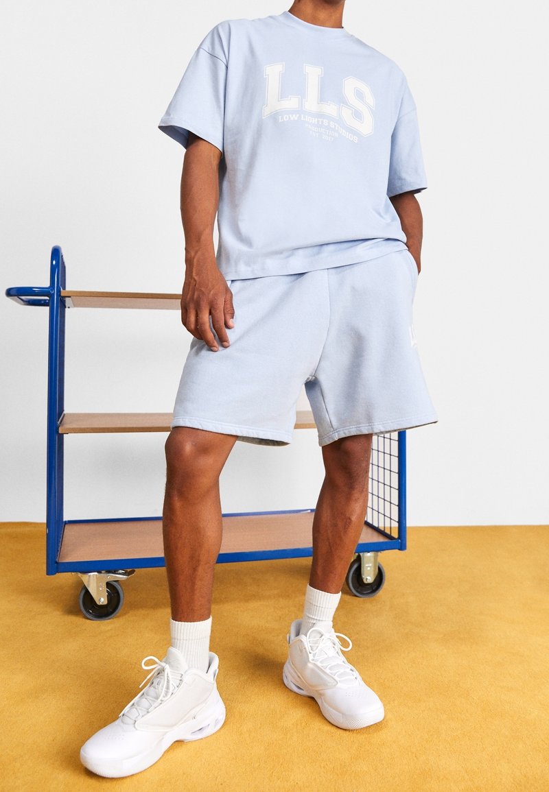 Personne portant un short LLS bleu clair et un T-shirt assorti, des chaussettes blanches et des baskets blanches, debout près d'un chariot bleu et en bois sur un sol jaune.