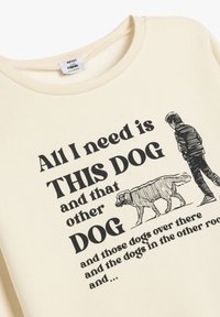 Sudadera color crema con impresión negra. Presenta una ilustración de un perro y el texto: "Todo lo que necesito es ESTE PERRO y ese otro PERRO."