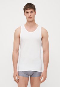 Weißes Baumwoll-Tanktop mit runden Ausschnitt, mit glatter Oberfläche und kontrastierenden Abschlüssen, getragen mit gestreifter grauer Unterwäsche.