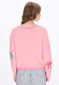 Rosa flauschiger Pullover mit langen Ärmeln, der auf den Ärmeln gestickte Schleifen in Grün, Lila und Rot sowie einen kontrastierenden pinken Saum aufweist.