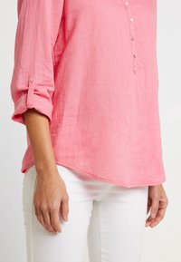 Camicia di lino rosa con apertura a bottoni, maniche arrotolate e orlo curvo, abbinata a jeans bianchi. Tessuto morbido e vestibilità rilassata.