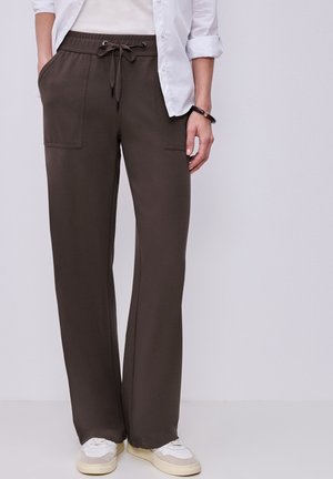 Osoba nosící tmavě hnědé kalhoty se šňůrkou v pase a předními kapsami, bílé tenisky, bílé tričko a černý náramek na pravém zápěstí.