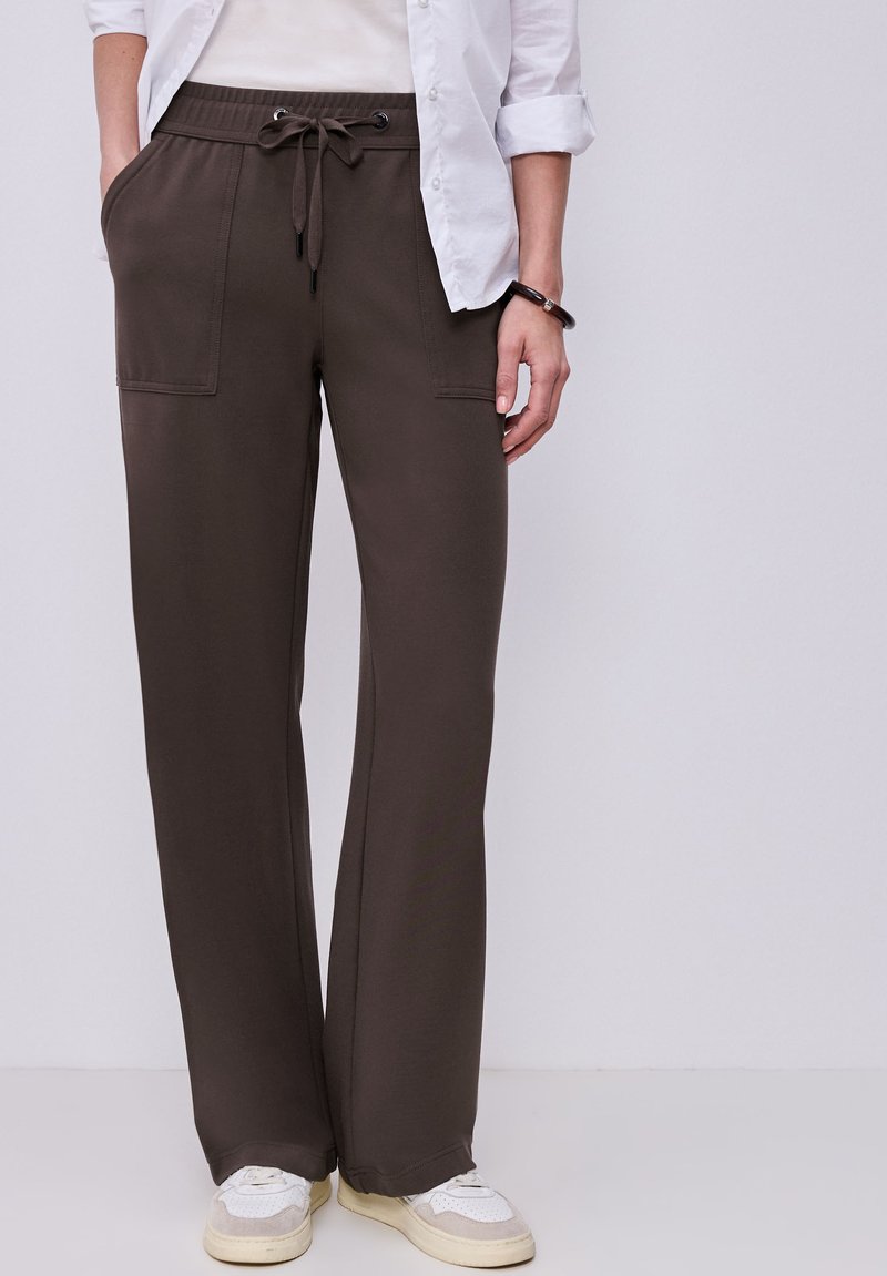 Persona que lleva pantalones marrón oscuro con cordón, bolsillos frontales, zapatillas blancas, una camisa blanca y una pulsera negra en la muñeca derecha.