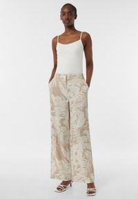 Brede broek in beige stof met witte bladmotieven, voorzien van zijzakken en een losse pasvorm. Model gecombineerd met witte sandaaltjes met bandjes.