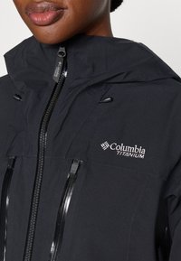 Juodas Columbia Titanium džemperis su kapišonu, turintis užtrauktukais užsegamas kišenes, vandeniui atsparų paviršių ir atspindinčius detales.