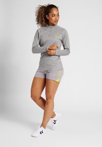 Maglia sportiva grigia chiaro a maniche lunghe con colletto a mezza zip e logo verde. Abbinata a pantaloni corti grigi con dettagli gialli e sneaker bianche.