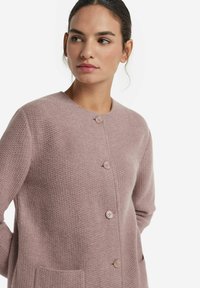 Kobieta ubrana w teksturowany sweter w kolorze przykurzonego różu z czterema guzikami i kieszeniami naszywanymi, patrzy w lewo na tle białym.