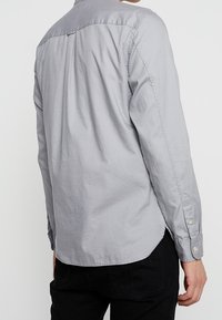 Mężczyzna w jasnoszarej, długiej koszuli z długim rękawem i czarnych spodniach, widziany plecami na jednolitym tle.