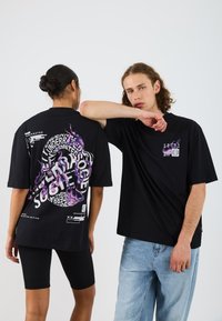 Černé oversized trička s odvážnými fialovými grafickými designy na zádech a přední straně, nošená s černými šortkami a světle modrými džíny.