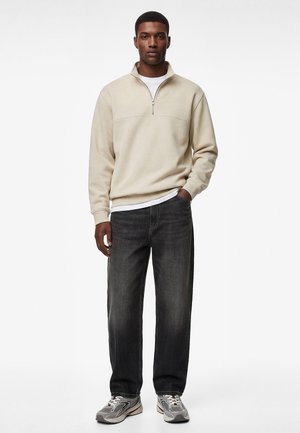 Homme portant un sweat-shirt beige à fermeture éclair quart, un sous-vêtement blanc, un jean gris foncé, des baskets grises et blanches, debout avec une main dans la poche.