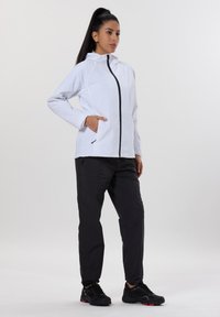 Valkoinen kevyt takki, jossa on musta vetoketju ja taskut, yhdistetty mustiin housuihin ja mustiin kenkiin, suunniteltu ulkoiluun.