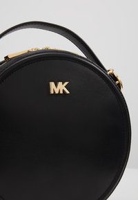 Kulatá černá kožená kabelka se zlatým logem MK, zlatým zipem a krátkým uchem na jednobarevném světlém pozadí.