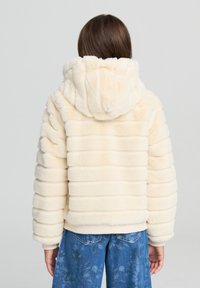 Futrzana kurtka w kremowym kolorze z poziomym pikowaniem, dużym kapturem i ściągaczem u dołu. W zestawie z niebieskimi dżinsami w kwiatowy wzór.