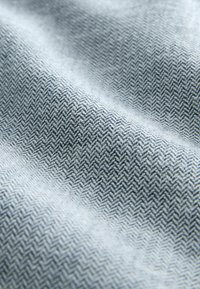 Stoff in Fischgrätmuster in hellblauen und grauen Tönen, mit einer glatten Textur und einem dezenten Glanz. Besitzt diagonale Linien und feine Details.