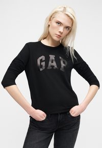 Νέα γυναίκα με ξανθά μαλλιά, ντυμένη με μαύρο μακρυμάνικο μπλουζάκι GAP και μαύρο τζιν, όρθια με τα χέρια στις τσέπες μπροστά σε απλό φόντο.