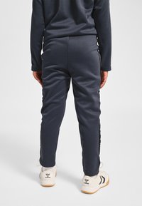 Granatowe spodnie sportowe o gładkiej fakturze, z czarnymi paskami po bokach i białymi akcentami w postaci logo. Połączone z beżowymi tenisówkami.