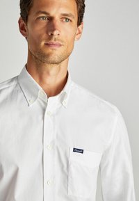 Camicia bianca con bottoni, realizzata in cotone. Presenta un colletto a punta, una tasca frontale con un'etichetta blu e una texture sottile. Design semplice.