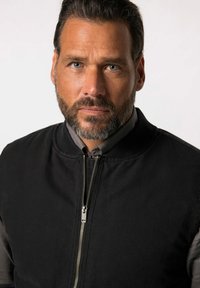 Chaqueta bomber negra hecha de un tejido duradero, con cremallera frontal, cuello acanalado y una textura suave. Incluye una camiseta gris debajo.