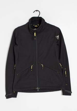 Veste softshell noire avec fermeture éclair sur toute la longueur, col montant et tirettes de fermeture éclair jaunes, exposée sur un cintre contre un fond blanc.
