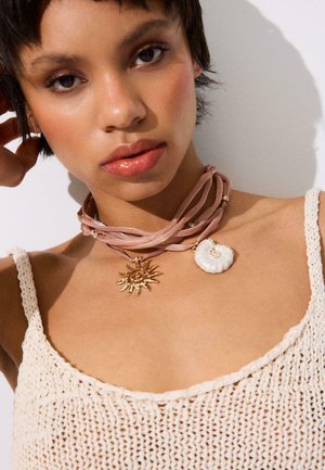 Vrouw draagt een beige gehaakte tanktop en een lichtroze chokerketting met gouden zon- en witte schelpenhangers tegen een witte achtergrond.