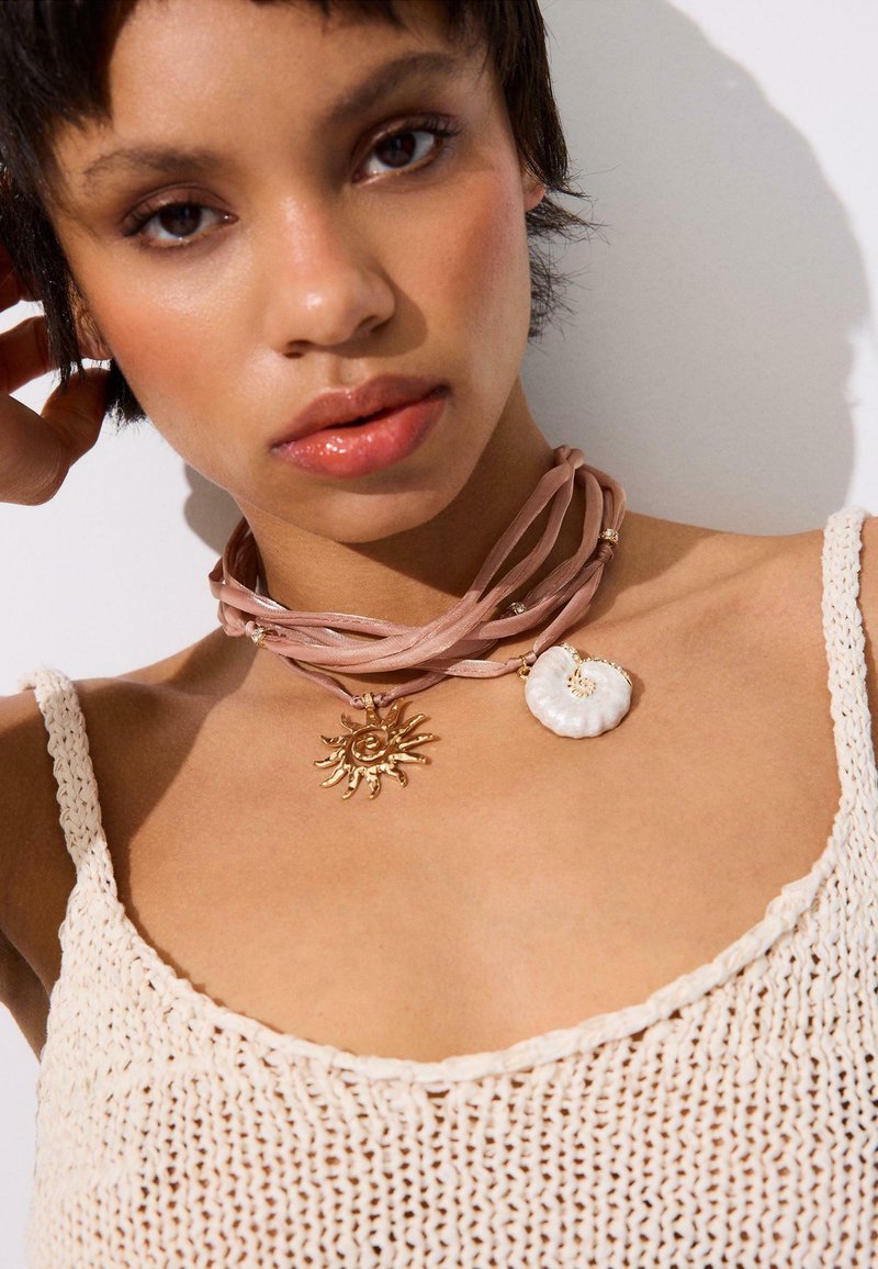 Femme portant un débardeur en crochet couleur nude et un collier ras-du-cou rose clair avec des pendentifs soleil doré et coquillage blanc sur un fond blanc.