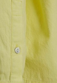 Tejido de camisa en un amarillo claro con un acabado texturizado, que presenta un botón plateado y un detalle de bolsillo parche cerca de la tapeta de los botones.