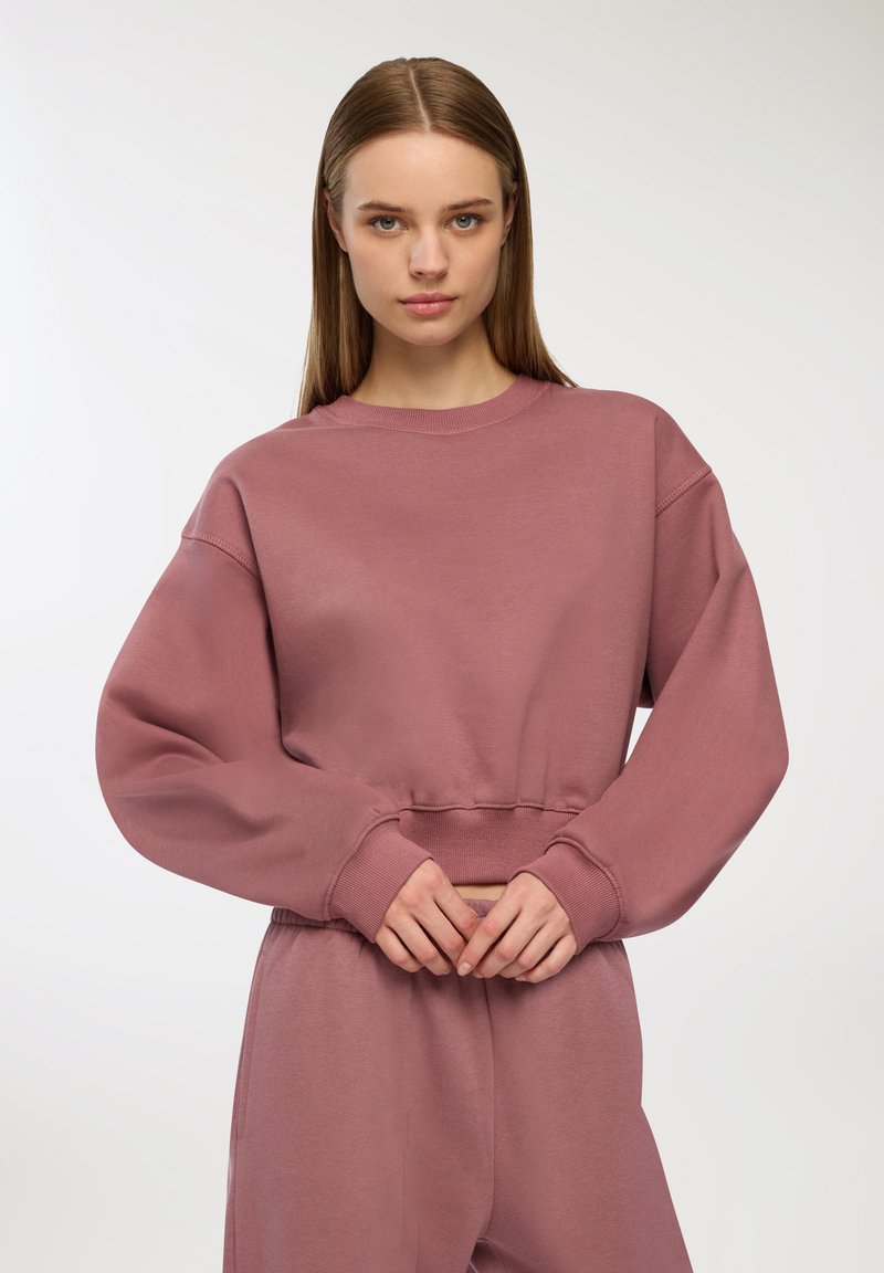 Felpa cropped in rosa polveroso, realizzata in tessuto morbido. Presenta spalle scese e polsini a coste, con una vestibilità rilassata e un design minimale.