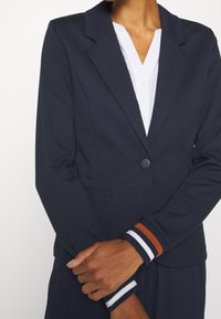 Blazer bleu marine avec un col cranté, une fermeture à un bouton et une coupe structurée. Les poignets présentent des rayures avec des accents marron. Texture de tissu lisse.