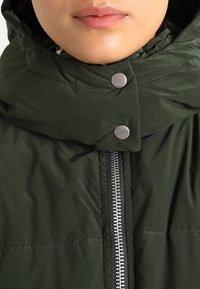 Ciemnozielona kurtka puchowa z wysokim kołnierzem, wyposażona w srebrne zatrzaski i kontrastujący zamek błyskawiczny. Teksturowana tkanina z poziomymi liniami.