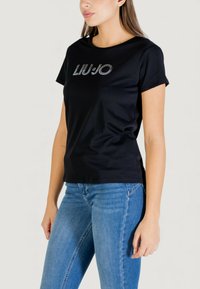 T-shirt noir à manches courtes avec un col rond et le logo « LIU JO » en strass argentés sur le devant. Associé à un jean bleu.