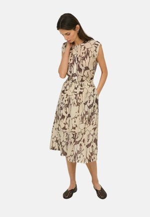 Mujer con cabello oscuro que lleva un vestido midi floral sin mangas beige y marrón con bolsillos, de pie y mirando hacia abajo con una mano cerca de su rostro.