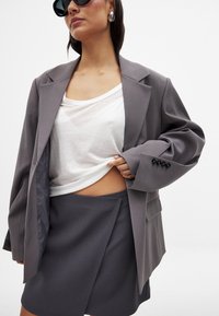 Blazer gris surdimensionné avec des poignets boutonnés, associé à un haut transparent blanc et une jupe portefeuille grise. Tissu lisse et lignes épurées. Lunettes de soleil noires portées.