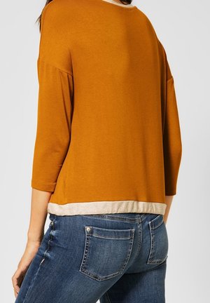 Femme portant un haut à manches longues jaune moutarde avec un ourlet beige et un jean en denim bleu, vue de dos et de la taille vers le haut.