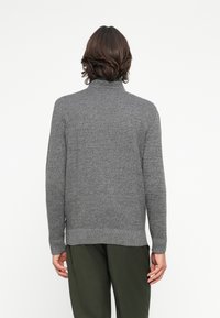 Maglione grigio a collo alto lavorato a maglia con una finitura testurizzata, bordi a coste sui polsini e sull'orlo, e una vestibilità rilassata. Indossato sopra pantaloni verde scuro.