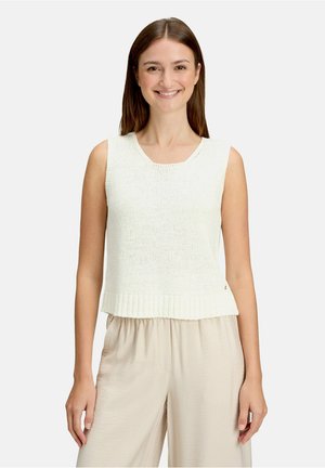Jeune femme souriante, portant un haut sans manches en maille blanche et un pantalon beige ample, debout devant un fond clair uni.