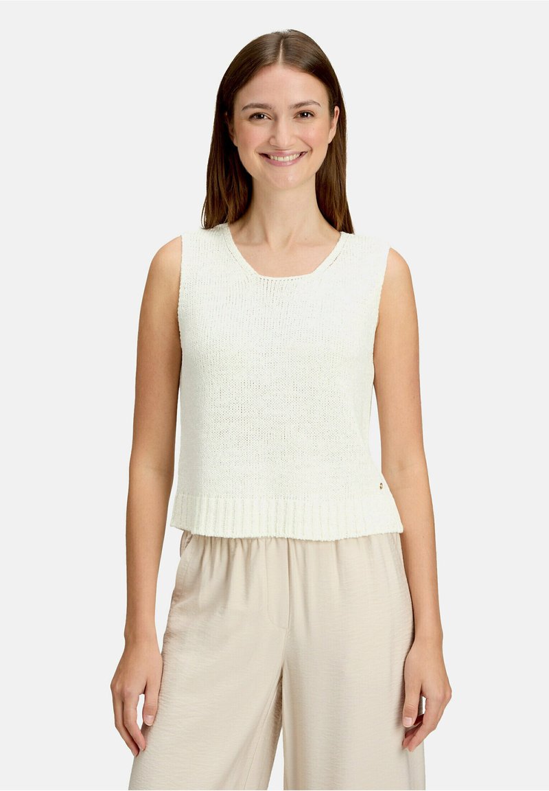 Jeune femme souriante, portant un haut sans manches en maille blanche et un pantalon beige ample, debout devant un fond clair uni.