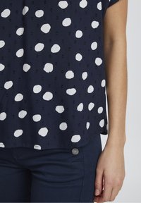Navy blue top featuring white polka dots, short sleeves, a relaxed fit, and textured fabric with a small, visible button accent.