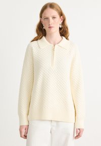 Maglione color crema con un motivo a nido d'ape texturizzato, dotato di colletto con zip e polsini e orlo a coste.