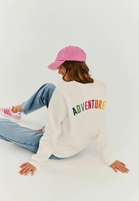 Bílá mikina s barevným nápisem "ADVENTURE" na zádech, kombinovaná se světle modrými džíny a růžovou baseballovou čepicí, doplněná o růžové sportovní boty.