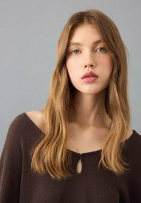 Bruine gebreide trui met een off-shoulder ontwerp en een sleutelgatknopdetail bij de halslijn. Zachte textuur en natuurlijk haar dat het gezicht omlijst.