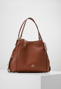 Hnědá kožená kabelka s texturovaným povrchem, zlatými kovovými akcenty, dvojitými uchy a štítkem s logem. Uvolněný design, zaoblený tvar.