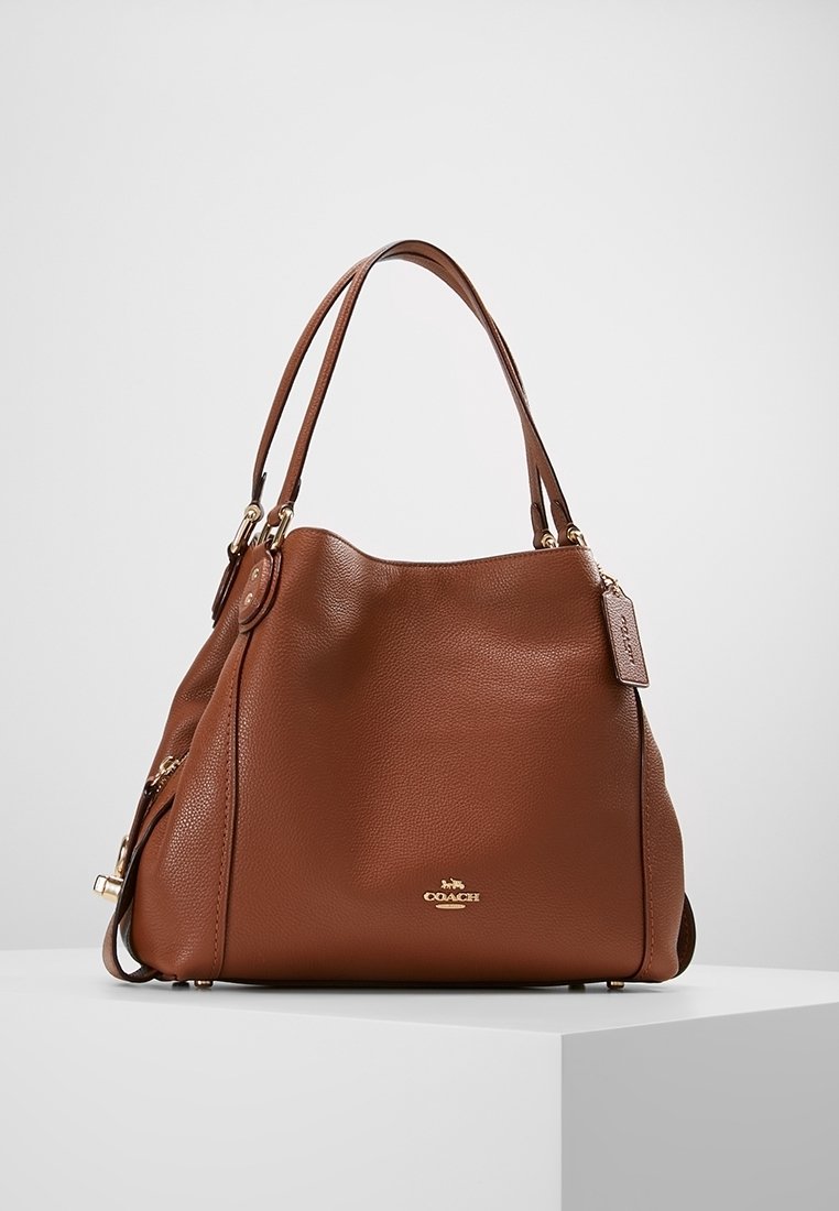 Hnědá kožená kabelka s texturovaným povrchem, zlatými kovovými akcenty, dvojitými uchy a štítkem s logem. Uvolněný design, zaoblený tvar.