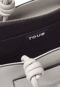 Sac à main en cuir gris avec une finition texturée. Dispose d'une bandoulière torsadée et d'un intérieur noir avec un logo "TOUS" visible à l'intérieur. Fermeture éclair.