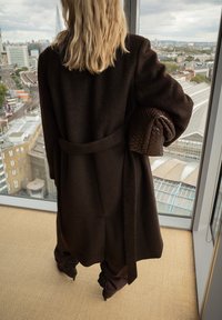 Brązowy wełniany płaszcz z paskiem, teksturowana tkanina i przesadnie duże rękawy, trzymany z plecioną, ciemno-brązową torbą. Widok na miasto przez okno.